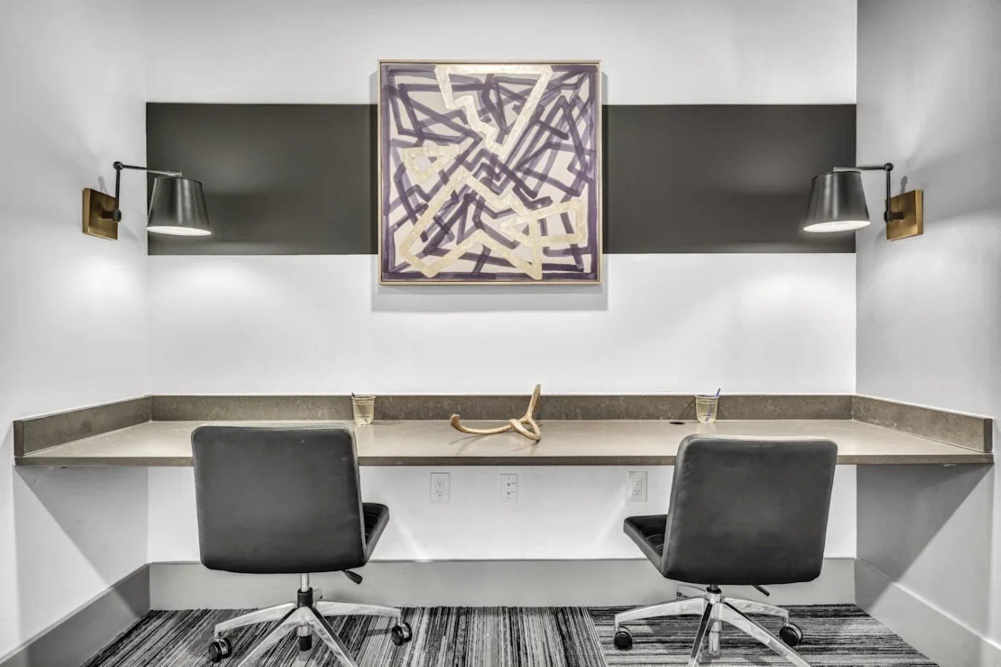 A co-working study space featuring a built-in double desk with black rolling chairs, wall sconces, and abstract art.